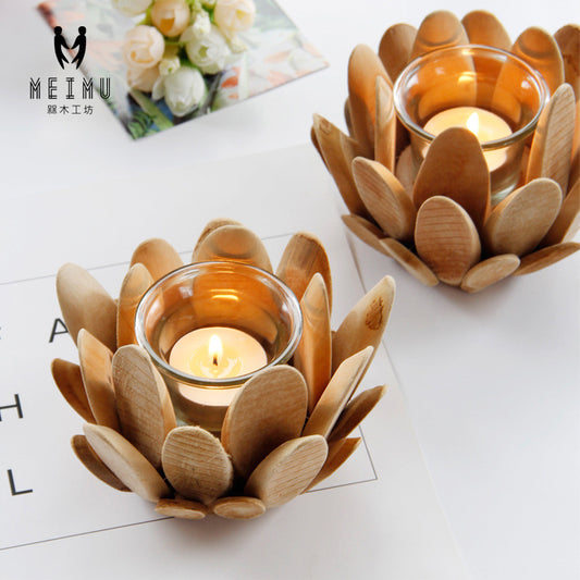 Two wooden candlestick holders, shaped like flowers, with candles lit inside them, placed on a table with a floral decoration in the background.